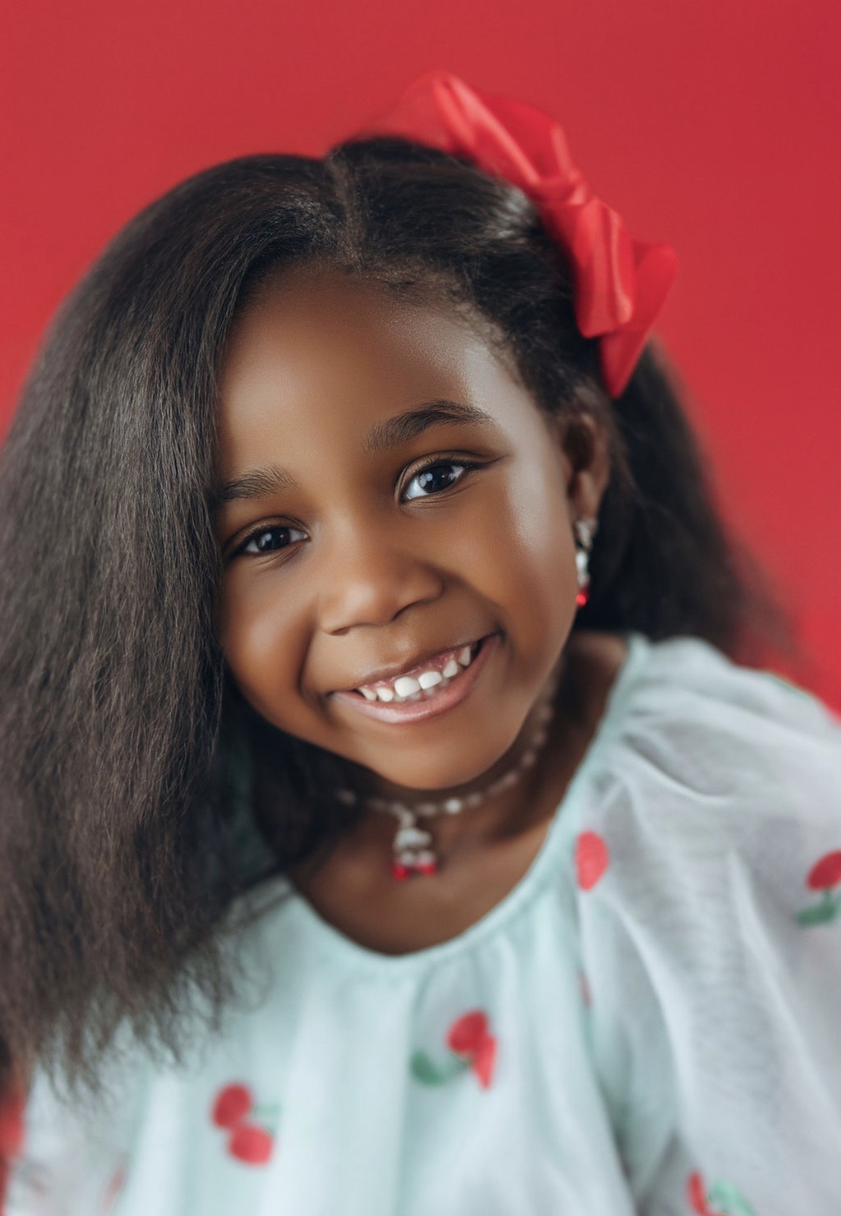 Noe smiling boldly, red bow, cherry dress on bold red backdrop